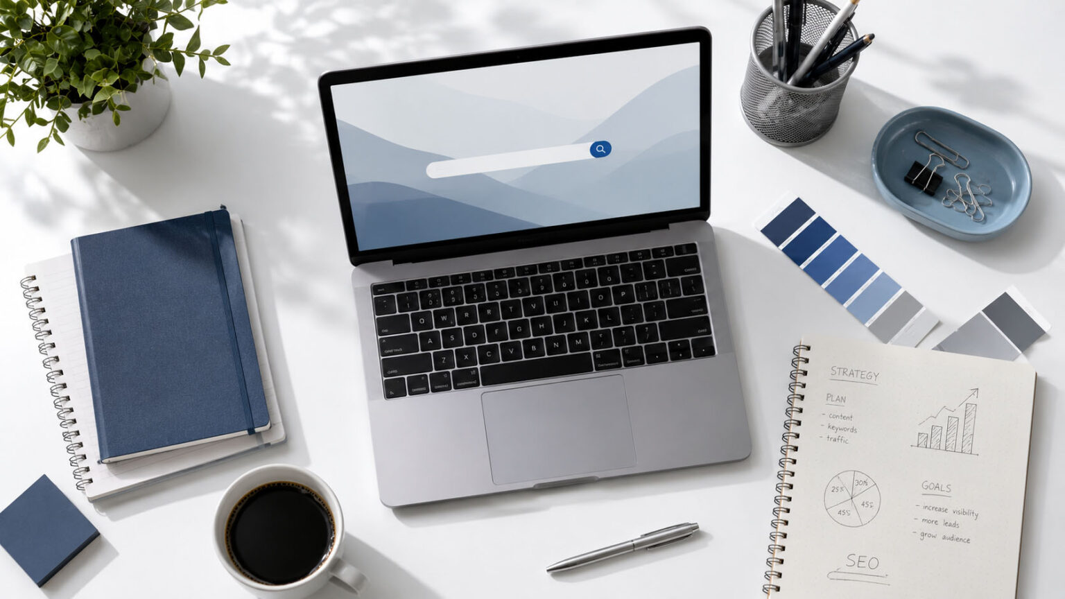Overhead view of a modern workspace: laptop on a white desk surrounded by notebooks, color swatches, a pencil cup, and a cup of coffee.